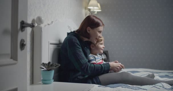 Happy mother and son playing video games on bed at home. Boy holding wireless controller in hands, woman teaching how to play. Family having fun.  - Materiał filmowy, wideo