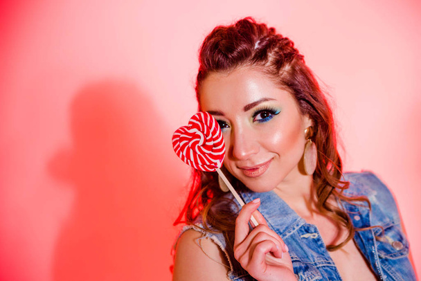 Beautiful brunette girl with blue makeup, pigtails and a sleeveless denim vest posing on a red background with a heart-shaped lollipop d - Fotoğraf, Görsel