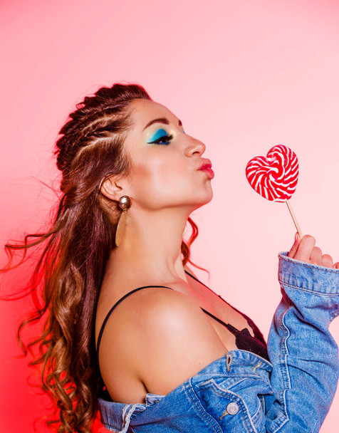 Beautiful brunette girl with blue makeup, pigtails and a sleeveless denim vest posing on a red background with a heart-shaped lollipop d - Photo, image