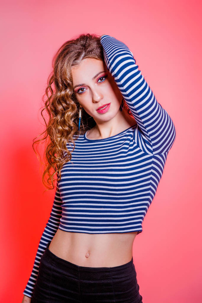 Happy beautiful brunette girl with red makeup, curly hair and a blue stripes sweater posing against a red background - Photo, image