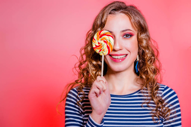 Happy beautiful brunette girl with red makeup, curly hair and a blue stripes sweater posing against a red background. One eye is covered with candy - Foto, Imagen