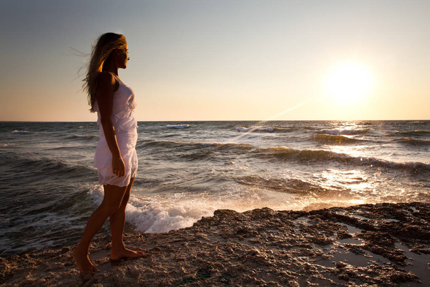 Young smiling blond woman in white dress standing on rocks and looking at sunset over wavy sea water - Fotoğraf, Görsel