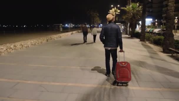 Un viajero con una maleta roja vino a descansar, caminando por la noche a lo largo del paseo marítimo - Imágenes, Vídeo