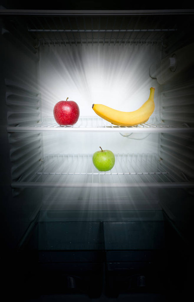 Conceptual creative photo of fresh red and green apples and banana glowing in the dark in an open empty refrigerator. Weight loss diet concept. - Foto, imagen