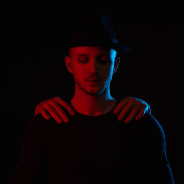 Ηρεμία νεαρός όμορφος άνδρας με μαύρο καπέλο κοιτάζει κάτω, αρσενικά χέρια τον κρατούν από τους ώμους, κόκκινο και μπλε φως πέφτει πάνω του, απομονώνονται σε μαύρο φόντο - Φωτογραφία, εικόνα