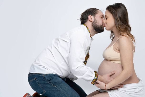Coppia giovane seduta a terra, uomo barbuto in camicia nazionale bianca che bacia la sua bella moglie bruna incinta, ritratto in studio
 - Foto, immagini
