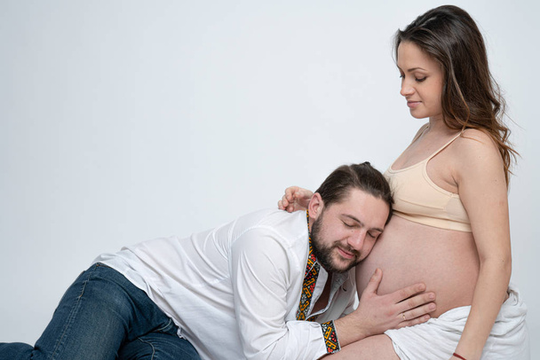 Bonito homem de camisa branca está ouvindo com os olhos fechados para sua bela barriga grávida esposa, isolado sobre fundo branco
 - Foto, Imagem