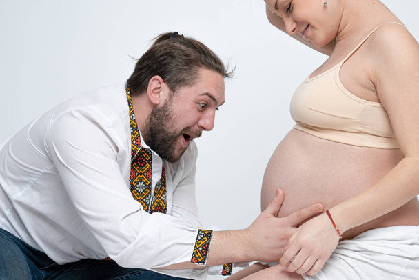 Χαρούμενος γενειοφόρος νεαρός που αγγίζει την κοιλιά της όμορφης γυναίκας του. Έγκυοι σύζυγοι νιώθουν αγάπη. Λευκό φόντο - Φωτογραφία, εικόνα
