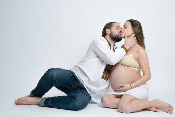 Fiatal pár ül a földön, szakállas férfi fehér nemzeti ing csók gyönyörű terhes barna felesége, stúdió portré - Fotó, kép
