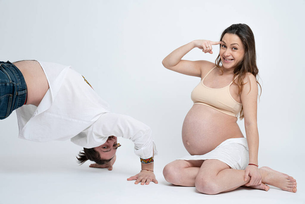 Sorrindo morena grávida mulher em sutiã bege sentado no chão, olhando para a câmera e torce um dedo perto da cabeça, enquanto seu marido, vestido com camisa branca, fica na posição de ponte
 - Foto, Imagem