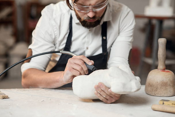 Attentive stonemason with stylish beard in protective goggles detailing creative sculpture of woman torso at his workplace. - Фото, зображення
