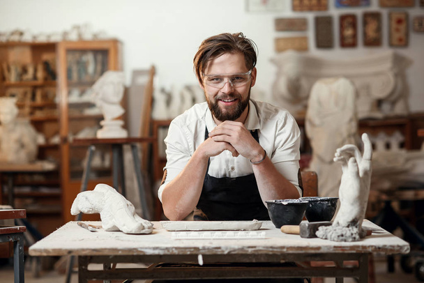 Portrait of attractive joyful bearded male sculptor which sitting on the workshop interior background and looks at the camera with smile. - Valokuva, kuva