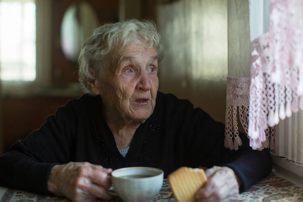 Μια ηλικιωμένη κυρία πίνει τσάι με μπισκότα στο σπίτι..  - Φωτογραφία, εικόνα