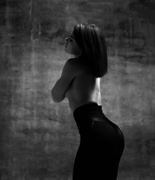 Young slim brunette woman in black sport shorts standing backwards with naked back over dark wall background - 写真・画像