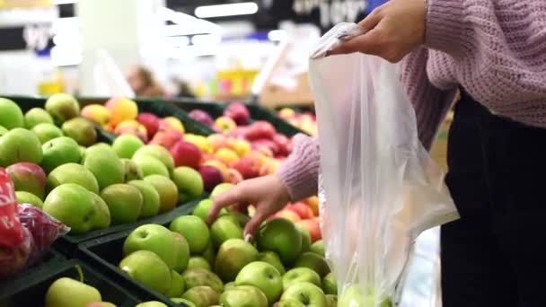 Una guapa chica toma manzanas verdes y las coloca en una bolsa de plástico. Comprar alimentos para una nutrición adecuada y proporcionar vitaminas al cuerpo. Mujer seleccionando manzanas frescas en la venta de supermercados de tiendas de comestibles - Imágenes, Vídeo