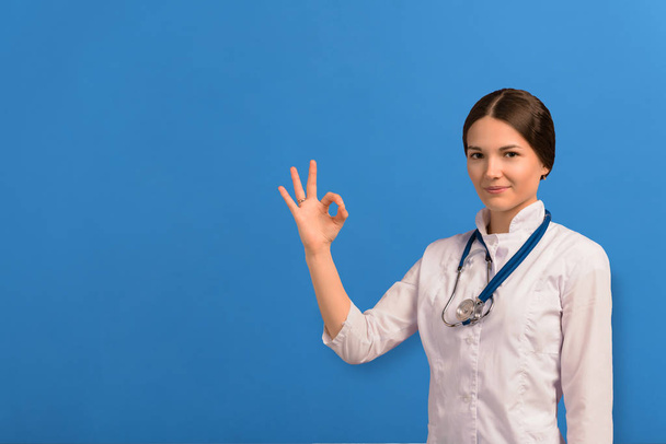 Portrait of beautiful doctor girl in white coat with stethoscope showing OK sign looking at camera and smiling isolated on blue background - Foto, imagen