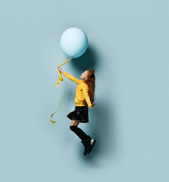 Teenage female in yellow sweatshirt, black skirt, knee-highs, boots. She holding balloon, posing sideways on blue background. - Fotoğraf, Görsel
