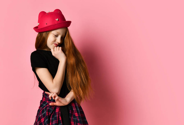 Teenager in black dress, checkered shirt on waist, red hat. She thought, touching her face, posing on pink background. Close up - Photo, image