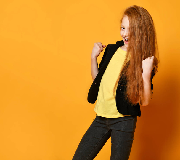 Ginger schoolgirl in black jacket, pants, yellow t-shirt. She is smiling, looking satisfied, posing on orange background. Close up - Foto, afbeelding