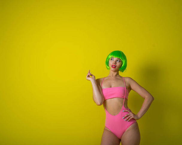 Beautiful woman in a green short wig and pink bikini posing on a yellow background. Portrait of a girl with sensual red lips. - Photo, Image