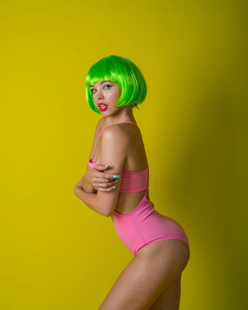 Beautiful woman in a green short wig and pink bikini posing on a yellow background. Portrait of a girl with sensual red lips. - Fotografie, Obrázek