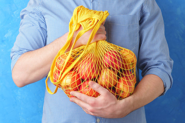Un hombre con una camisa azul sostiene una bolsa de cuerdas con manzanas rojas en sus manos
. - Foto, Imagen