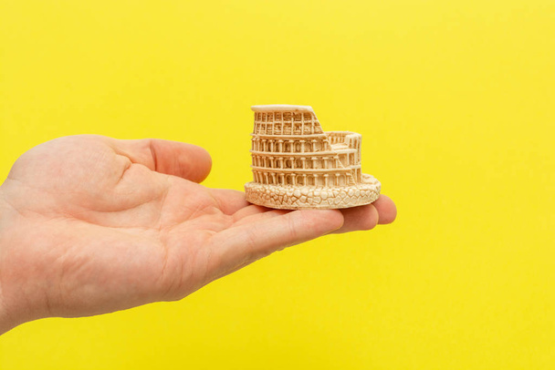 Male hand holds a coliseum figurine on a yellow background. Travel Italy or study Italian language concept - Foto, Imagem