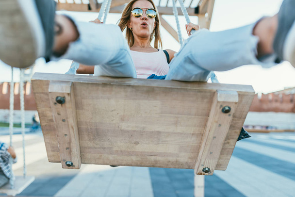 long-haired beautiful young blonde in a pink T-shirt, sunglasses, light blue jeans and tied at the hips denim jacket, swinging on a swing and smiles. Selective focus macro shot with shallow DOF - Foto, Bild