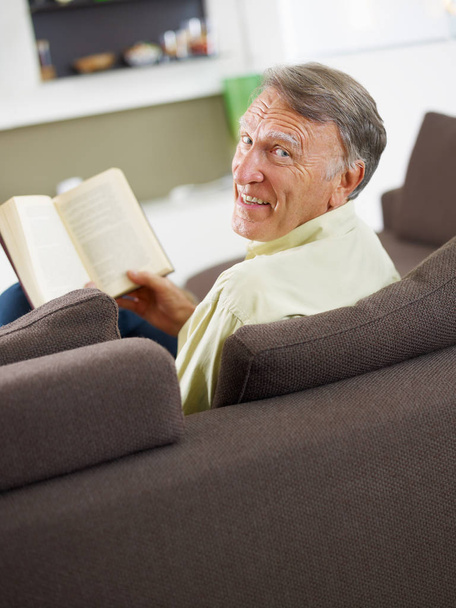 senior man reading book at home and looking over shoulders  - Фото, зображення