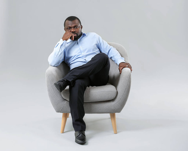 Afro-Amerikaanse man ontspannen in fauteuil tegen grijze achtergrond - Foto, afbeelding