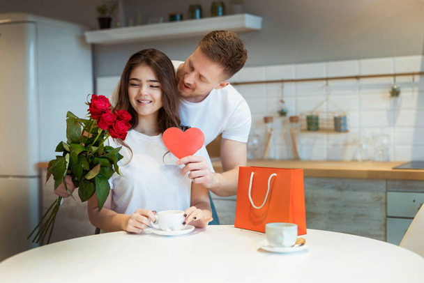 mladý pohledný manžel překvapuje svou milovanou ženu s růžemi a dárek na svatého Valentýna během ranní kávy v kuchyni, šťastný romantický výročí - Fotografie, Obrázek