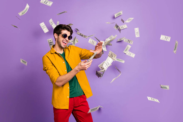 Photo of white cheerful brown haired trendy casual man with bristle throwing cash with crazy emotions on face in red pants yellow shirt isolated violet pastel color background - Fotografie, Obrázek