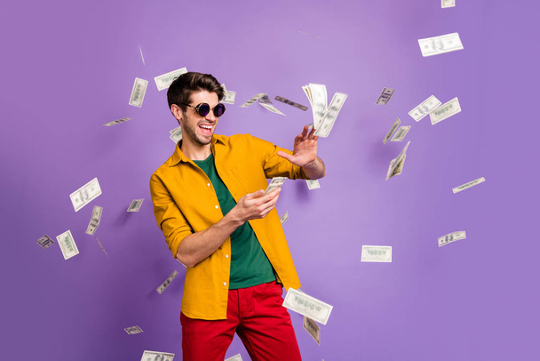Photo of cheerful excited white trendy man throwing cash away showing his wealthiness smiling toothily in red pants isolated violet color background - Foto, Imagen