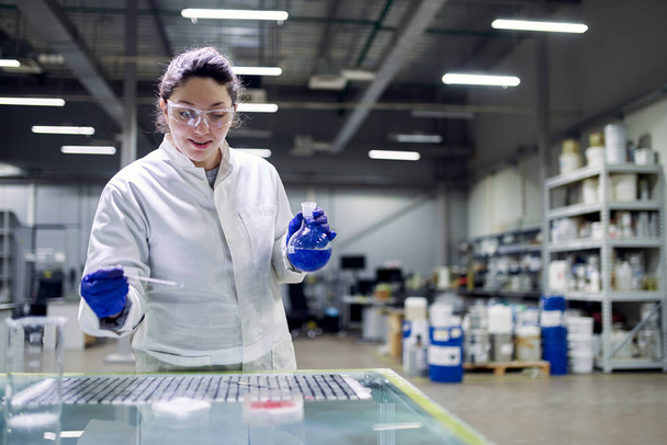 Laboratorium brunette vrouw met fles met blauwe vloeistof in haar handen controleert kwaliteit - Foto, afbeelding