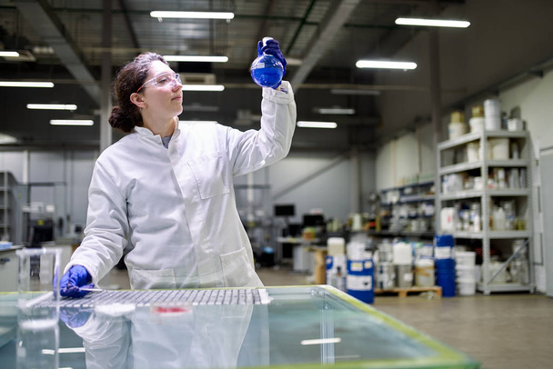 Laboratorní bruneta žena v bílém laboratorním plášti s baňkou s modrou tekutinou v rukou kontroluje kvalitu - Fotografie, Obrázek