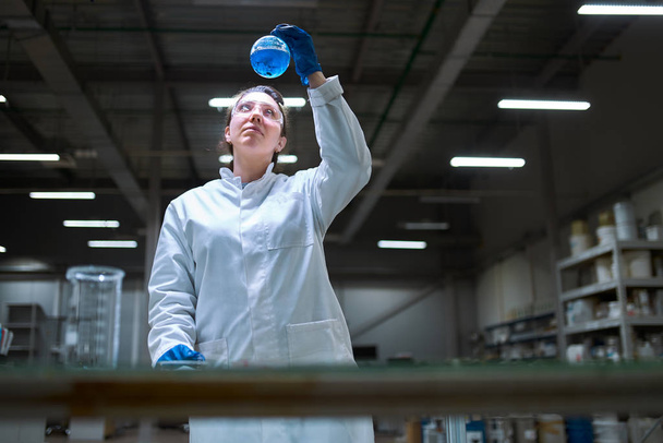 Young lab woman in white coat with flask with blue liquid in her hands checks quality on defocused background - Фото, изображение