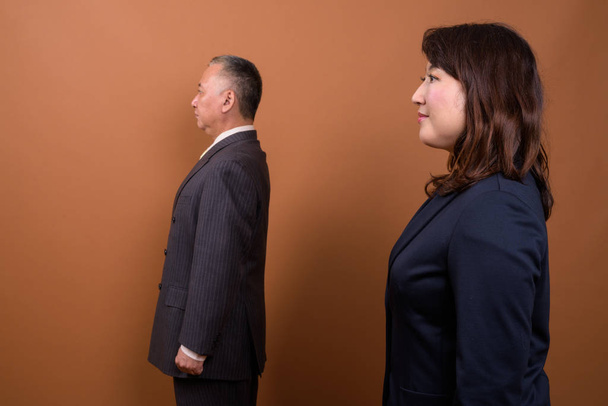 Studio shot of mature Japanese businessman and mature Japanese businesswoman together against brown background - Фото, зображення