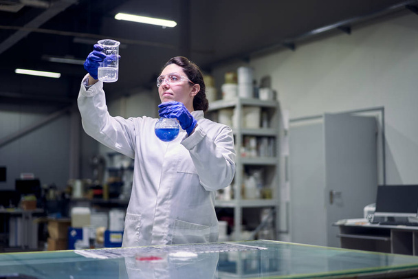 Brunette lab girl in glasses and white coat with flask with blue liquid in her hands conducts experiments - Фото, изображение