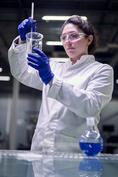 Young lab girl in glasses and white coat with experimental glass in her hands conducts experiments - Фото, зображення