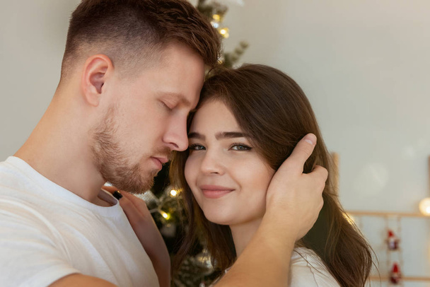 půvabný pár manžel objímající svou krásnou manželku stojící v blízkosti vánočního stromečku, jak se usmívají v místnosti zdobené na oslavu nového roku slavnostní nálada milostný příběh - Fotografie, Obrázek