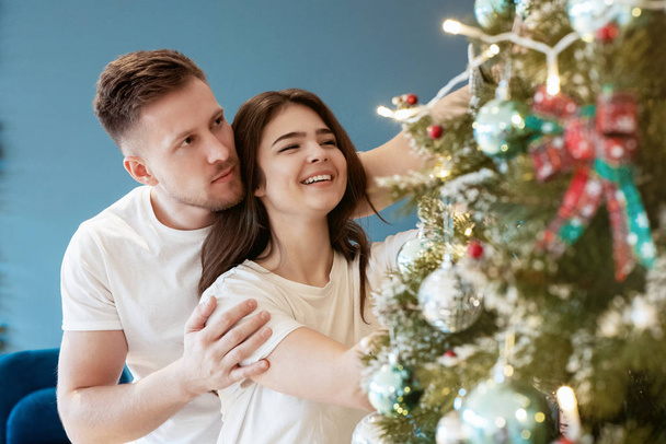 lovely couple husband hugging his beautiful wife decorating together christmas tree both smiling in room decorated for celebrating new year festive mood love story - Photo, image