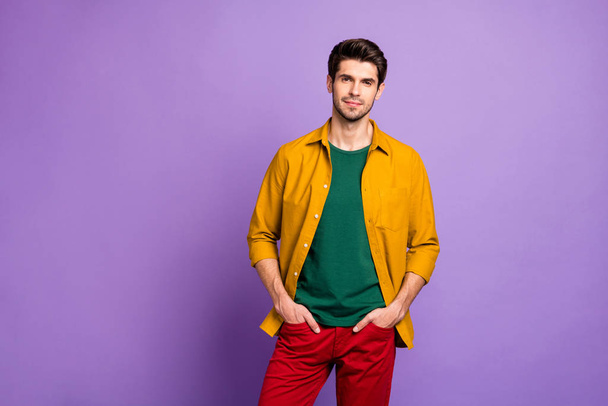 Photo of confident serious man standing with hands in pockets of red pants looking at you isolated violet pastel color background - Фото, изображение