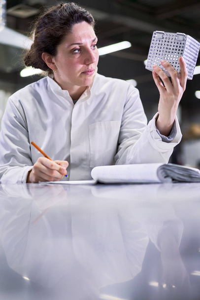 Brunette woman in white coat with metal cube sits on table in laboratory - Fotografie, Obrázek