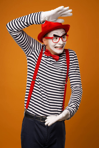 Surprised mime with white face in red hat and striped t-shirt on blank orange background - Фото, изображение