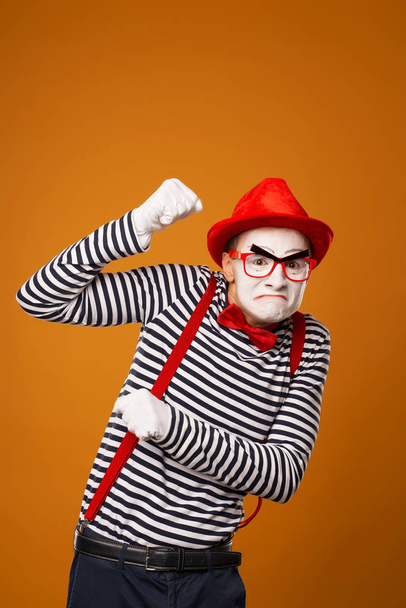 Sad mime with white face in red hat and striped t-shirt on blank orange background - Фото, зображення
