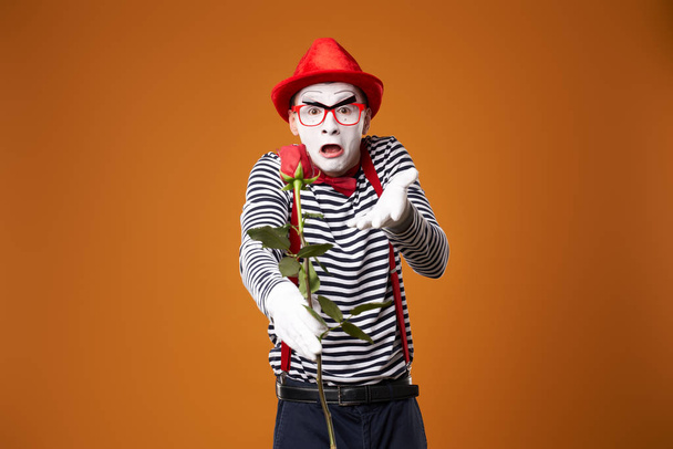 Sad mime man in red hat and vest with rose in hands on orange background - Photo, Image
