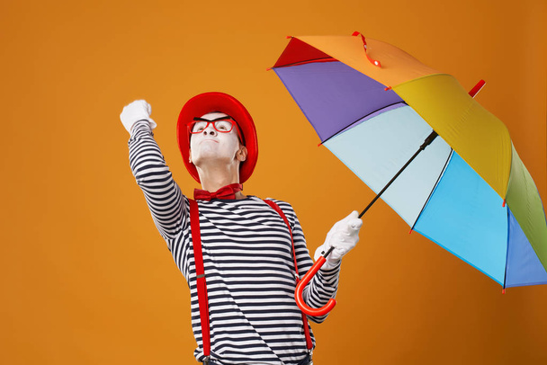 Mime looking up with multi-colored umbrella in hand and showing fist isolated on blank orange background - Fotografie, Obrázek