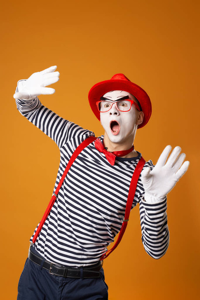 Young mime male in white gloves and red hat looking at side on orange background - Фото, зображення