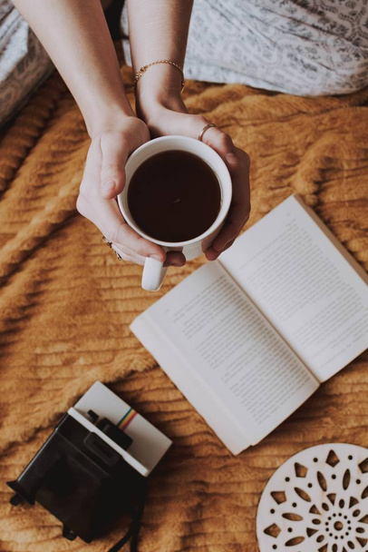 A closeup shot of a woman holding a cup of coffee in her hands next to a book and other objects on a smooth surface - Foto, Bild