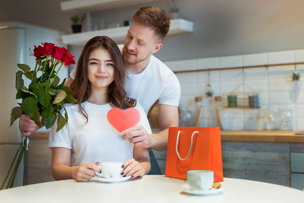 young handsome man surprising his beloved wife with roses and gift on saint valentine's day during breakfast coffee in the kitchen, happy romantic anniversary - Photo, Image